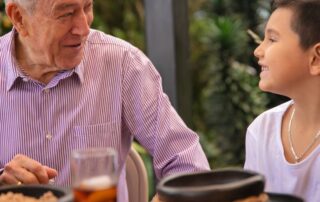 An elderly man and a young boy smile at each other while sitting at a table outdoors, sharing a warm moment together. Plates of food and a glass of iced drink are visible on the table in front of them.